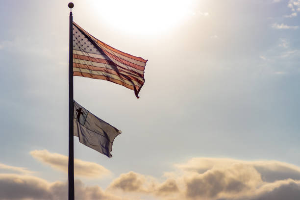 An American and Christian flag flutter in the wind atop a flagpole with the afternoon sun and golden colored clouds behind them.
