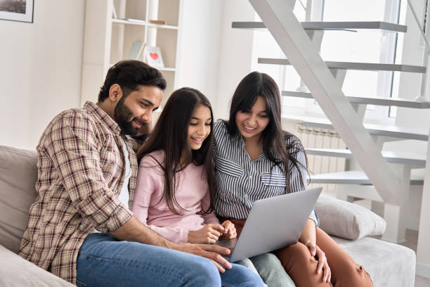 happy indian family couple with child daughter using laptop computer at home. smiling parents and teen kid bonding watching streaming online tv or doing ecommerce shopping together sitting on sofa. - apartment television family couple imagens e fotografias de stock
