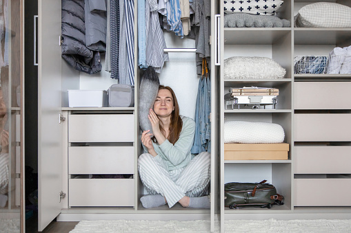 Smiling housewife in pajamas relaxing sleeping on pillow at wardrobe. Funny playful young woman hiding in modern cupboard full of clothes. Pleasant domestic female enjoying weekend at home