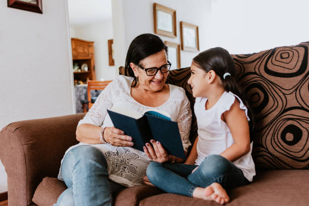 latin grandmother with granddaughter sitting on couch at home and reading book in mexico city - grandparent reading grandmother child imagens e fotografias de stock