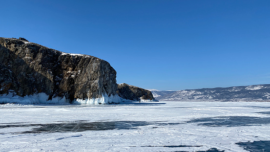 A beautiful winter landscape with majestic ice-covered rocks, mountains and hills. A tourist walks on the ice of frozen Lake Baikal.