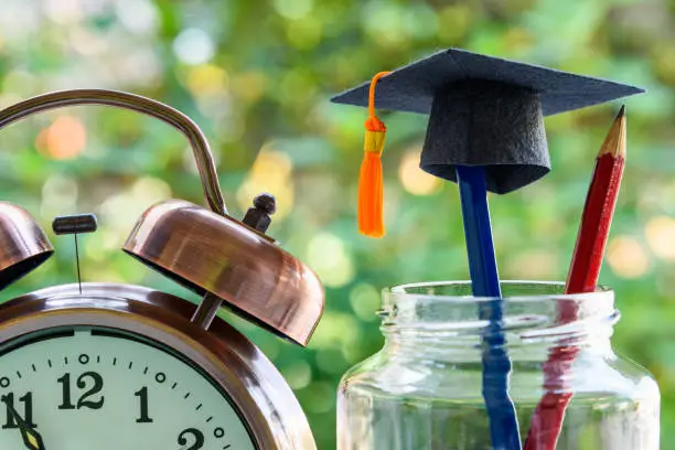 Photo of Black graduation cap or a mortarboard, a clock, a blue and a red pencils in a bottle