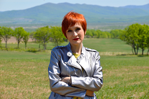 Portrait of Russian red-haired woman with short hair looking at the camera against the background of a field. Crossed arms