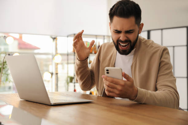 istock Emotional young man with smartphone at table in office. Online hate concept 1310223112