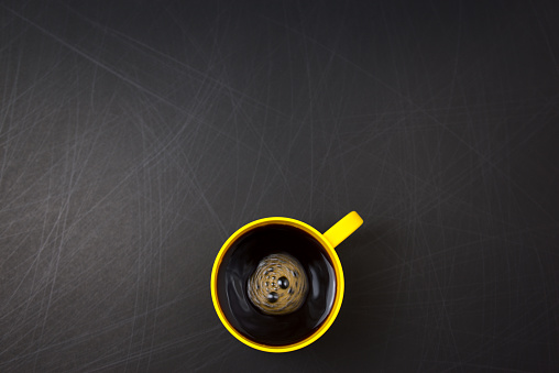 Clockwise stir. Yellow cup of black coffee flat lay on scrathed rough black background. Copy space for your text, image or message. Minimal, top view, horizontal image style. Off-center composition.