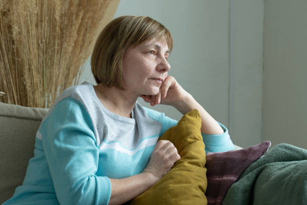 mujer anciana reflexiva se sienta en el sofá y mira por la ventana - pensive senior adult looking through window indoors fotografías e imágenes de stock