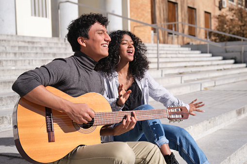 A young latin couple playing the guitar and singing sitting on stairs. Millennial generation.