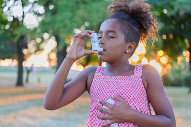 Beautiful Little African-American Girl is Having Asthmatic Problems Outside in Nature. Young Girl of African-American Ethnicity is Using Inhaler Due to the Problems with Asthma in the Public Park. asthmatic stock pictures, royalty-free photos & images