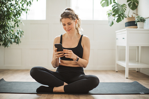 Mature woman practicing yoga at home in her living room. She is using phone to check social media.