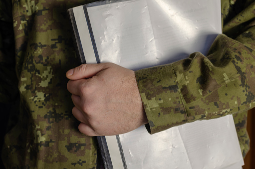 An officer is holding a plastic folder of documents. Black folder with documents inside against the background of a camouflage uniform. Adult middle-aged man. Selective Focus. Low key. Indoors.