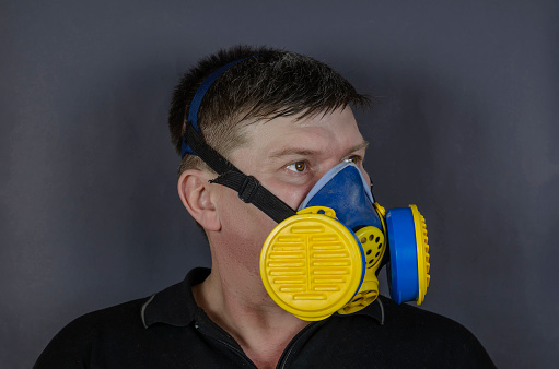 Portrait of a man in an industrial respirator on a gray background. An adult male with short hair wearing a blue and yellow respirator. Half mask with replaceable filters. Indoors. Safety first