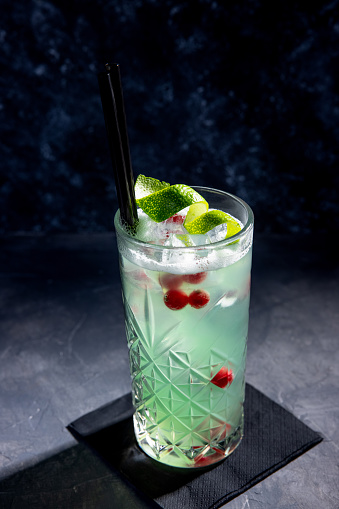 Mint colored cocktail on ice decorated with lime peel  and currant berries, black drinking straws and napkin as coaster, black background