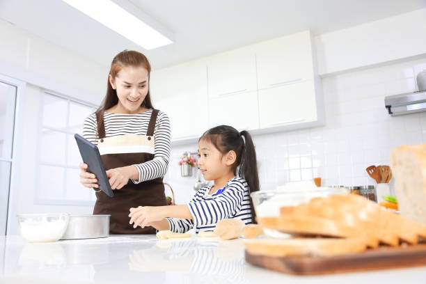 happy asian mother teaching her young daughter in baking bread from dough inside white modern kitchen while looking at cooking recipe - bun bread 7 grain bread dough imagens e fotografias de stock