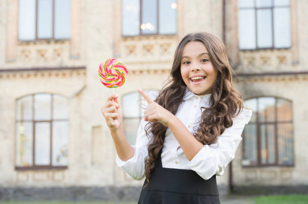 chica con el pelo rizado apuntando con el dedo en piruleta. dulces y dulces. niña feliz con uniforme elegante. de vuelta a la escuela. niño alumno en el patio de la escuela. calorías y alimentos útiles. niño en la pausa para el almuerzo - convenience holding sign teenager fotografías e imágenes de stock