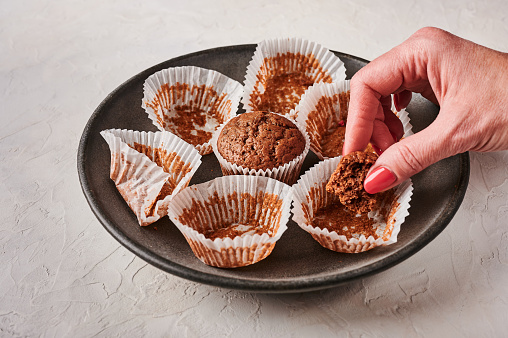 Woman's hand holds a piece of chocolate cupcake, next to empty wrappers on dark plate on wooden background, selective focus