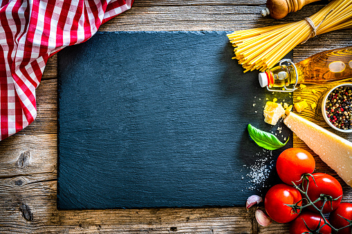 Italian food background: overhead view of uncooked spaghetti and ingredients for cooking a traditional Italian pasta arranged at the right of a wooden table making a frame and leaving useful copy space. The composition includes basil, fresh ripe tomatoes, olive oil, garlic, pepper, salt and Parmesan cheese. High resolution 42Mp studio digital capture taken with SONY A7rII and Zeiss Batis 40mm F2.0 CF lens