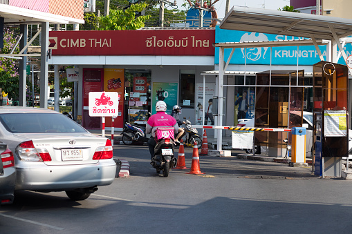 Thai food delivery person on motorcycle is crossing parking area in front of banks in Bangkok Ladprao. Man's fashion is pink colored for Food Panda company. He is passing exit gate of parking area, in background are some people at banks.