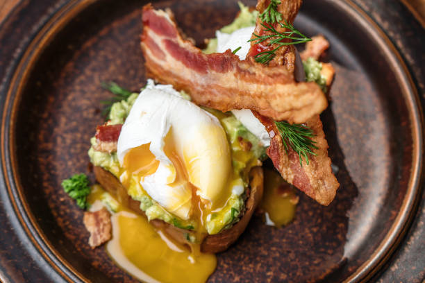 A serving of two Eggs Benedict, with  crispy ham, poached eggs and hollandaise sauce on a rustic brown plate