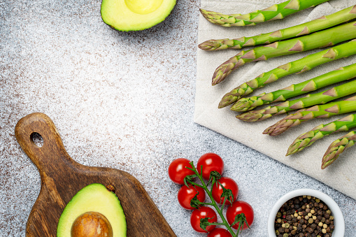 Healthy food concept. Keto diet. Fresh avocado on cutting board, asparagus on napkin, cherry tomatoes and pepper mix on textured grey background. Flat lay, copy space, top view.