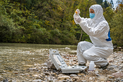 Female biologist in protective workwear adding reagent into a vial with water sample near river.