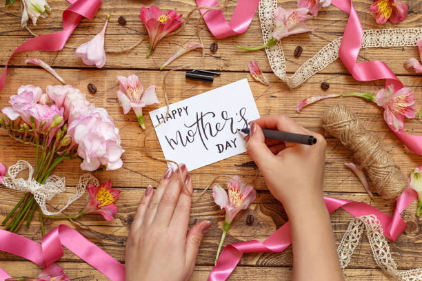 Hands Writing Happy Mother's Day card on a wooden table between pink flowers Hands Writing Happy Mother's Day card on a wooden table between pink flowers top view mother day handmade card stock pictures, royalty-free photos & images