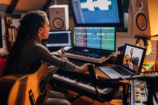 Modern young female artist with dreadlocks, an sound engineer working from her modern home office recording studio, while her female colleague helping her with advices during video call
