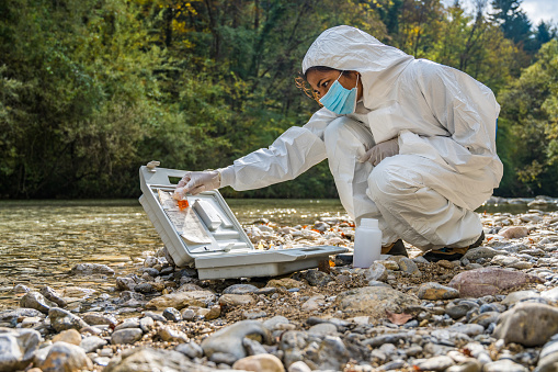 Female biologist in protective workwear examining water sample mixed with reagent in vial near river.