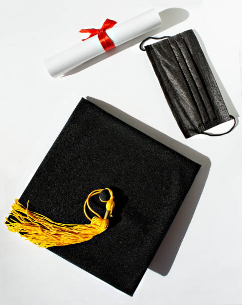 istock Black graduate cap with yellow tassel, paper scroll with red ribbon bow and protective face mask on white background 1310280848
