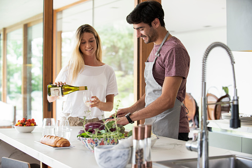 Smiling young man cutting vegetables and young woman pouring wine into glass in kitchen