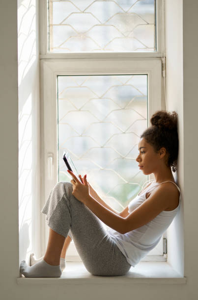 istock Side view of relaxed young woman reading a book using tablet pc while sitting by the window at home 1310283578
