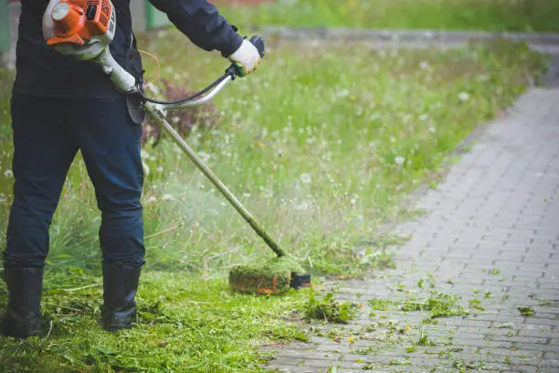 Photo of Worker in dark protective clothing with a gas mower in his hands, mowing grass in front of the house. A man mows grass with dandelions on a rainy spring day. Trimmer in the hands of a man.