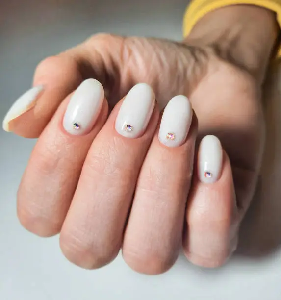 Gentle gel polish of milky color on short nails with sequins and rhinestones. Nude manicure with diamond and sequin design. Pastel color coating on the nails.