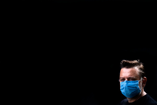 Portrait of mature man with protective blue color face mask . He is in front of black background.