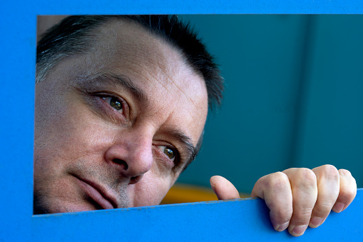 Extreme close up portrait of mature man looking though blue quadrilateral frame. The Photo was taken in outside. He is looking upward. He puts his hand on the frame. He seems melancholic and plaintive.