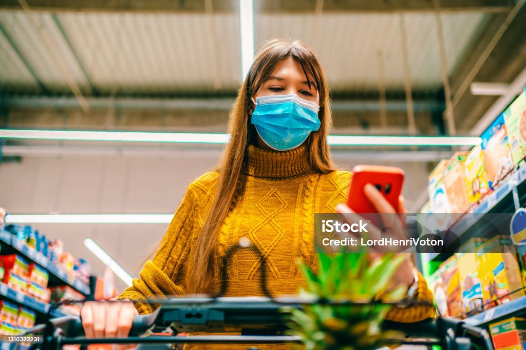 Woman checking shopping list on smartphone Young woman with face protective mask checking shopping list on smartphone at supermarket Protective Face Mask Stock Photo