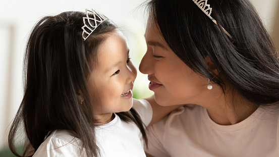 Close up loving Asian mother and little daughter wearing princess diadems enjoying tender moment, caring mum and adorable toddler child hugging cuddling, family having fun together