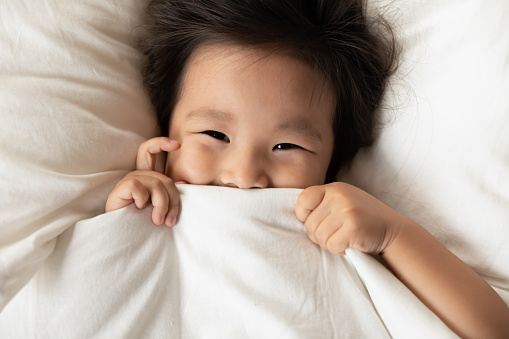 Top view close up funny portrait cute Asian little girl peeking from warm blanket, looking at camera, pretty adorable toddler child kid covering face with duvet, lying in cozy comfortable bed