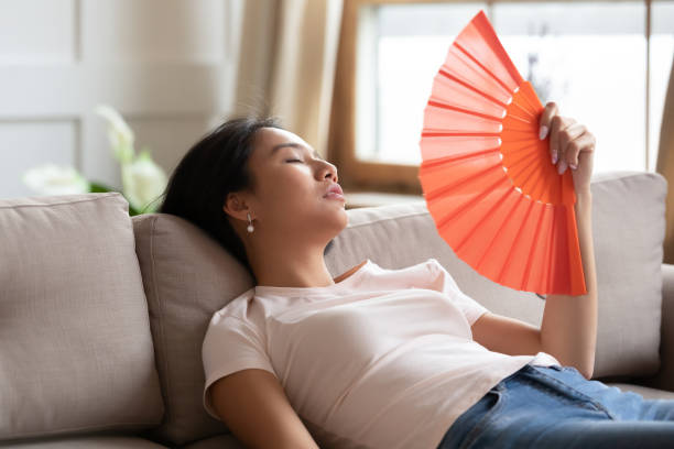 Overheated exhausted Asian woman waving paper fan, sitting on couch Overheated exhausted Asian woman waving orange paper fan, sitting on couch at home, tired girl feeling unwell, suffering from heating at home, feeling discomfort, hot summer weather or fever hot vietnamese women pictures stock pictures, royalty-free photos & images