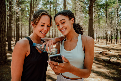 Two female friends texting on cellular device standing in forest after long run feeling good after exercise. High quality photo