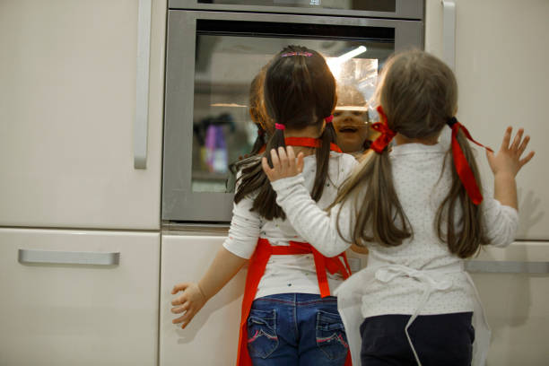 Rear view of two excited little girls standing in front of the oven in the kitchen and watching the gingerbread cookies they made being baked.