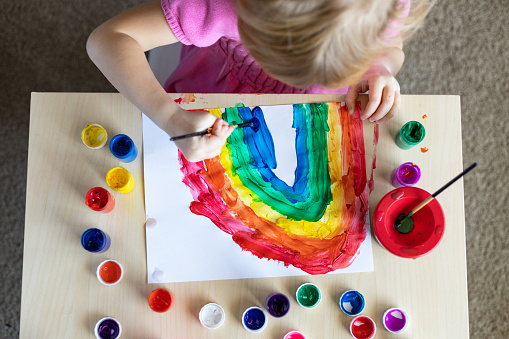 Little girl painting rainbow on paper with colorful paints sitting at the table at home