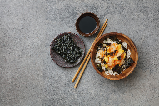Raw scallop rice bowl with mango, spicy mayo, tobiko caviar and nori chips. Flat lay top-down composition on concrete background.