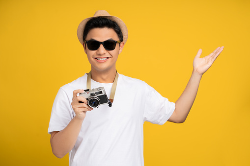 Happy young asian male tourists with open hand gestures and camera isolated on yellow background.