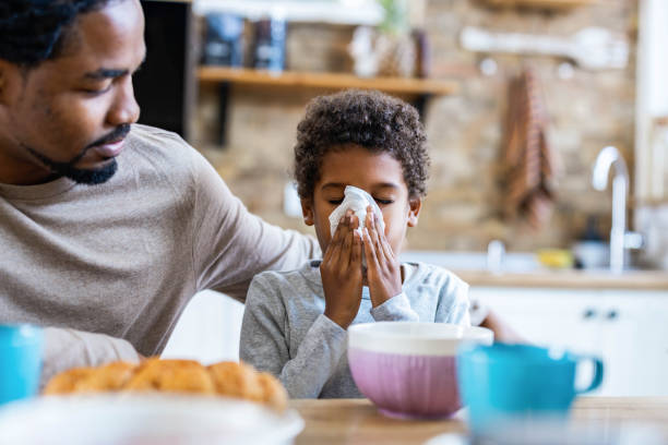 Single black father taking care of his sick son at home. Ill African American boy blowing his nose in dining room while his father is next to him. Focus is on boy. food allergies stock pictures, royalty-free photos & images