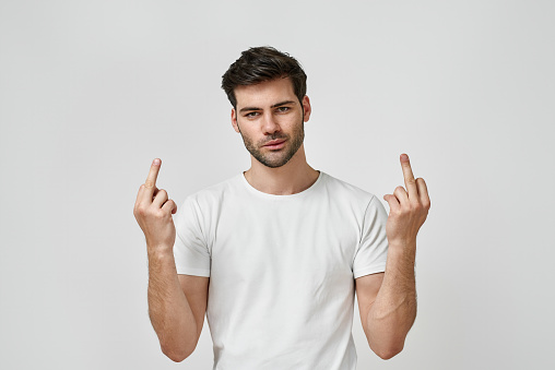 Handsome young bearded man wearing t-shirt showing middle finger doing fuck, provocation and rude attitude isolated on grey background. Hand gestures concept