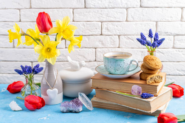 buquês de flores coloridas da primavera, xícara de café, livros, biscoitos, minerais de cristal de pedras preciosas sobre mesa azul no fundo de tijolos brancos. leitura e café da manhã. conceito de primavera, gemas, higge e interior aconchegante - hyacinth flower vase daffodil - fotografias e filmes do acervo