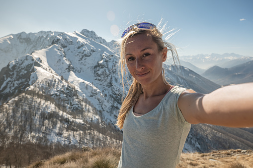 Cheerful young woman taking a selfie portrait on mountain top after hiking all the way up reaching summit