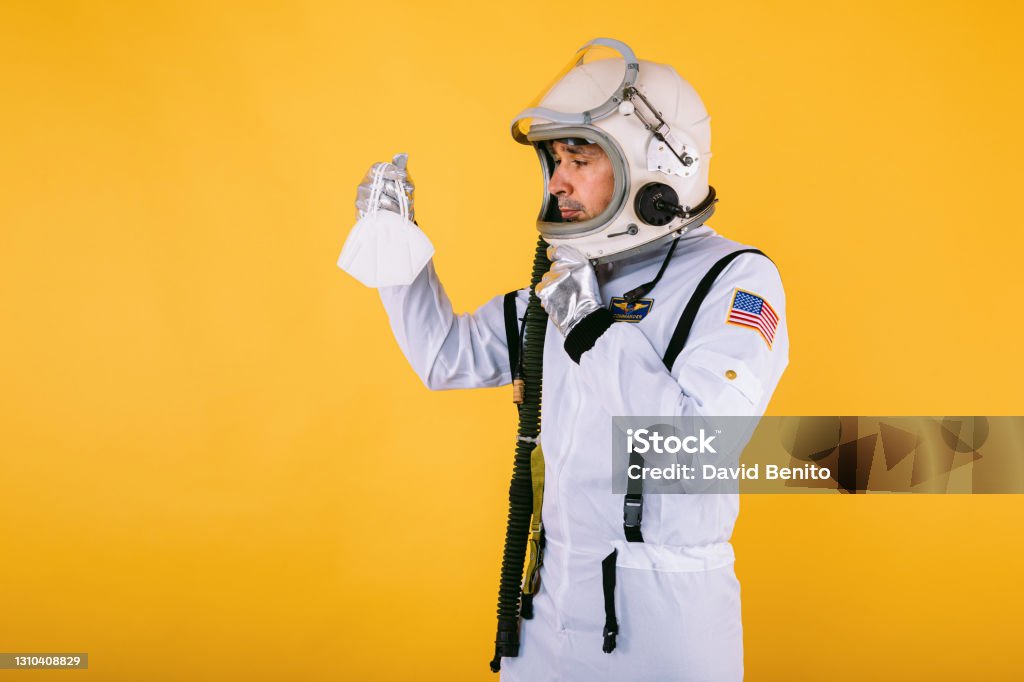 Male cosmonaut in spacesuit and helmet, holding a FPP2 mask, on yellow background. Covid-19 and virus concept Astronaut Stock Photo