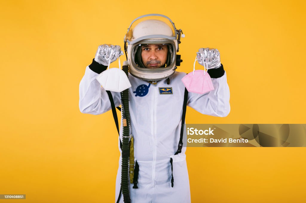 Male cosmonaut in spacesuit and helmet, holding two FPP2 masks, on yellow background. Covid-19 and virus concept 40-44 Years Stock Photo