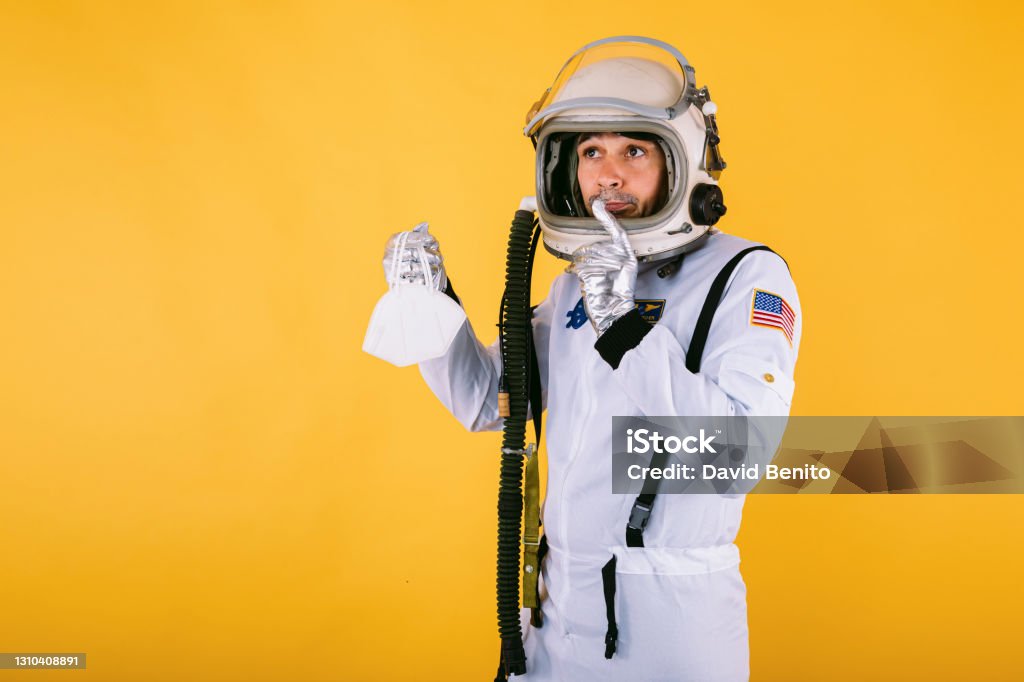 Male cosmonaut in spacesuit and helmet, holding a FPP2 mask, on yellow background. Covid-19 and virus concept 40-44 Years Stock Photo